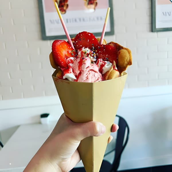 Hand holding a Kultura Cafe bubble waffle cone with strawberry ice cream, fresh strawberries, and sprinkles.