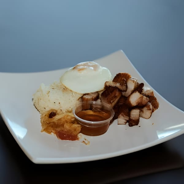 Filipino meal with crispy pork belly, fried egg, rice, and a side of sauce on a white plate.